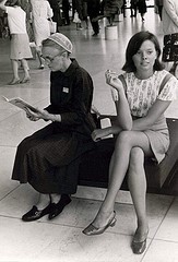 Old Mennonite女士坐在一位20歲、穿著迷你裙和高跟鞋、拿著香菸的年輕女子旁邊